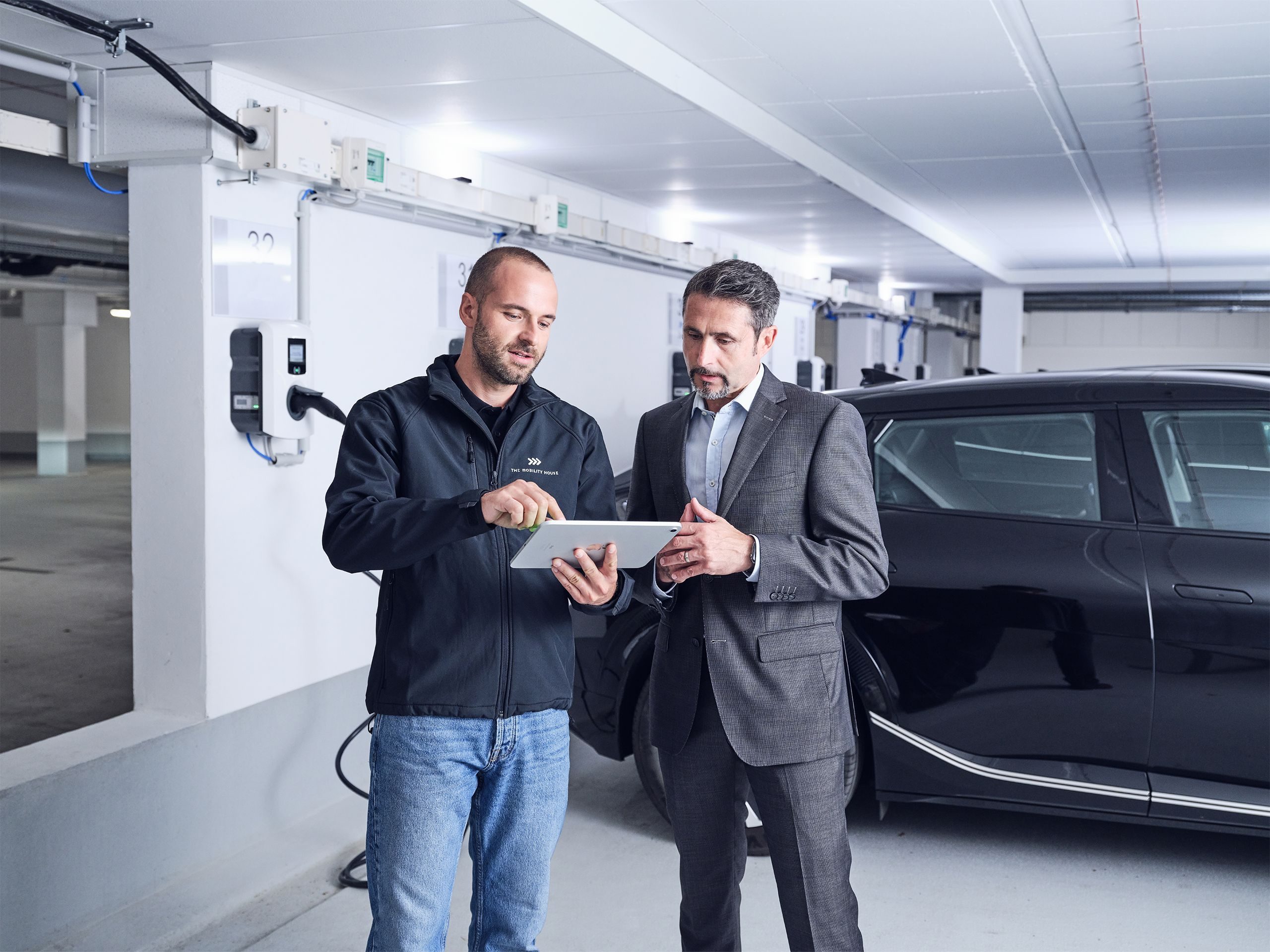 Elektriker und Kunde schauen gemeinsam auf Tablet in der Hand mit ladendem Elektroauto im Hintergrund